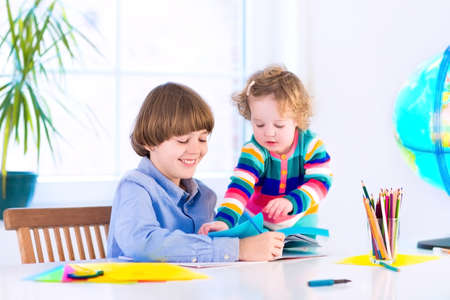 Two children, brother and sister, happy student boy and a little funny girl, doing homework together, reading books, cutting paper and drawing at a white desk with a globeの写真素材