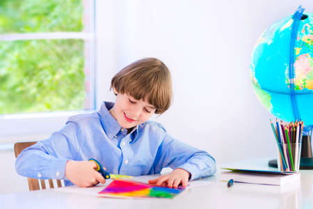 Happy smiling school boy, smart student, doing homework cutting paper, writing, drawing and reading a book at a white desk with a globe next to a window, back to school conceptの写真素材