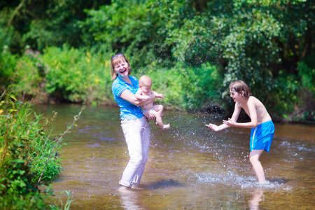 Active young woman playing with children, laughing happy boy and a cute little baby, splashing water and swimming in a river on a hot summer day hiking in th forestの写真素材