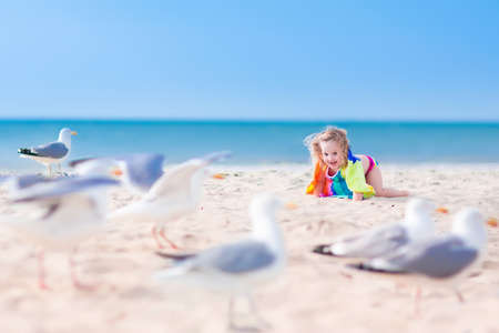 Funny lauging toddler, adorable little girl with curly hair in a colorful dress playing with seagull birds, running and jumping on a beautiful beach on a sunny hot summer dayの写真素材