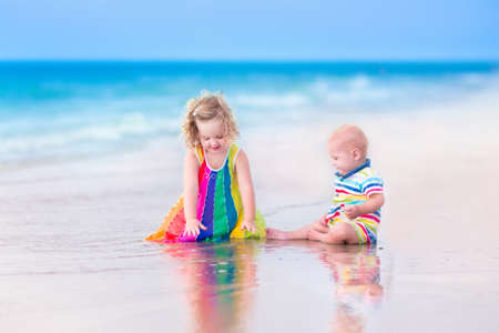 Two happy children, cute curly toddler girl and funny baby boy, brother and sister, playing together digging sand in water on a beautiful beach enjoying a hot summer vacation day on a tropical islandの写真素材