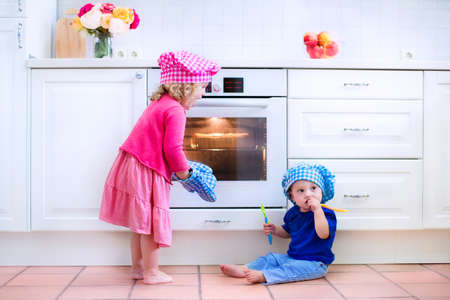 Cute kids, adorable little girl and funny baby boy wearing pink and blue chef hats playing with baking a pie in a sunny white kitchenの写真素材
