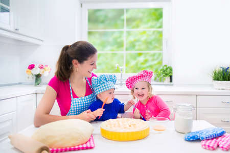 Young happy mother and her kids, adorable toddler girl and a little funny baby boy wearing pink and blue chef hats baking a pie together in a white sunny kitchen with big windowの写真素材