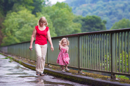 Young woman and cute laughing toddler girl walking on a bridge in a summer park after rainの写真素材