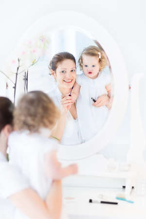 Young attractive woman and her cute funny toddler daughter wearing white dresses applying make up and cosmetics with a brush sitting in front of a round mirror in a white sunny bedroom  の写真素材