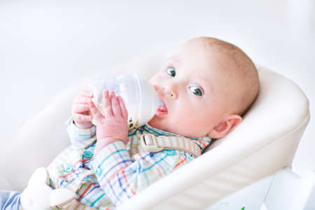 Adorable little boy sitting in a high chair drinking milk from a plastic bottle  