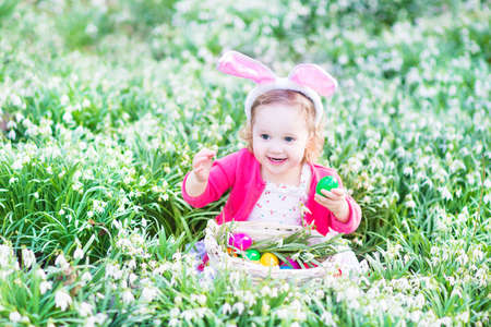 Adorable toddler girl wearing bunny ears playing with Easter eggs in a white basket sitting in a sunny garden with first white spring flowers  の写真素材