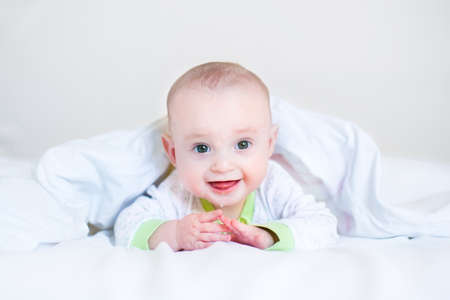 Adorable funny laughing baby playing peek-a-boo under a white blanket in a sunny bedroom waking up...