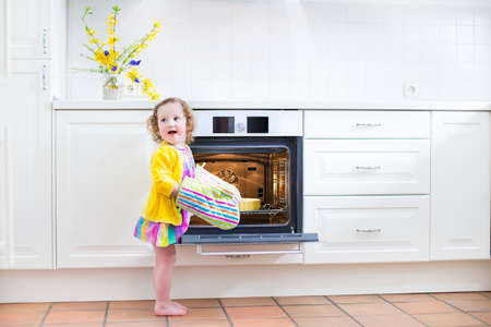 Adorable toddler girl in a yellow dress wearing colorful mittens playing in the kitchen next to a modern white oven helping by cooking and baking an apple pie in a home with white interiorの写真素材