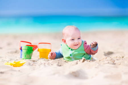 Adorable happy laughing little baby boy in a colorful sun protection suit playing with green, yellow and blue toy bucket and plastic shovel digging in sand on a beautiful exotic tropical beach with turquoise water の写真素材