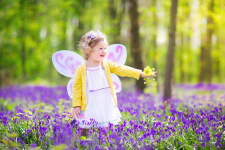 Adorable toddler girl with curly hair wearing a fairy costume with purple wings and yellow dress is...
