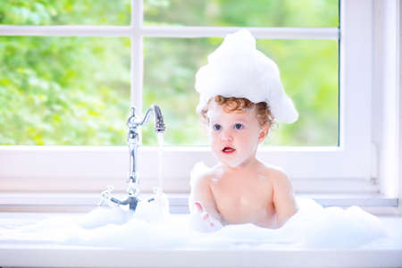 Funny little baby girl with wet curly hair taking a bath in a kitchen sink with lots of foam playing...