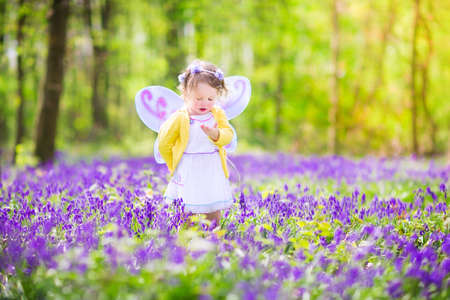 Adorable toddler girl with curly hair wearing a fairy costume with purple wings and yellow dress is...