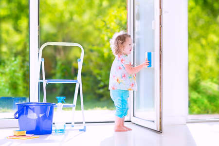 Cute laughing curly toddler girl washing a big window with a squeegee in beautiful white living room with door into the garden, standing on a ladder next to a blue bucket with water, detergent solution spray bottle and spongeの写真素材