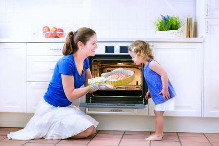 Young happy mother and her adorable curly toddler daughter wearing blue dress baking a pie together in an oven in a white sunny kitchen with modern appliances and devicesの写真素材