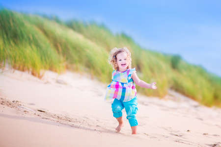 Happy funny little girl, adorable curly toddler, running and jumping in sand dunes enjoying family vacation at the North Sea, Holland, Netherlands on a hot sunny summer day at the beachの写真素材
