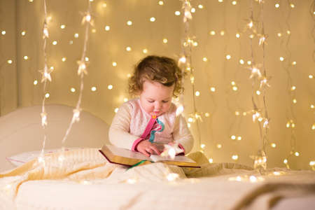 Cute little toddler girl reading a book in a dark room with Christmas lights の写真素材