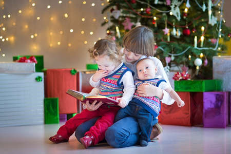 Portrait of three kids, smiling boy, toddler girl and a newborn baby boy, reading together under a beautiful Christmas tree の写真素材