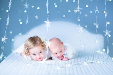 Adorable toddler girl and her newborn baby brother playing together between beautiful blue lights on a white bed の写真素材