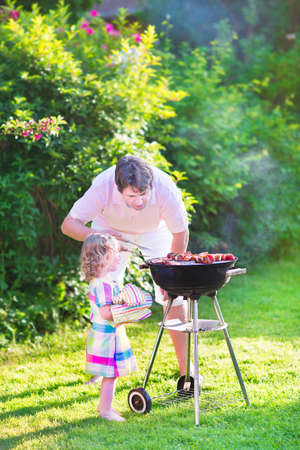 Young loving father and his cute happy toddler daughter, adorable curly girl wearing a colorful dress, having fun together grilling meat and vegetables in a sunny garden on a hot summer dayの写真素材