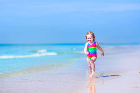 Happy laughing little girl in a colorful rainbow bathing suit runnig and playing on the ocean coast in water splashes on beautiful tropical island beach with turqouise clear water having fun on vacationの写真素材