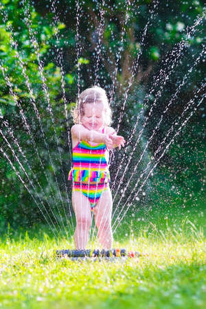 Funny laughing little girl in a colorful swimming suit running though garden sprinkler playing with water splashes having fun in the backyard on a sunny hot summer vacation day の写真素材