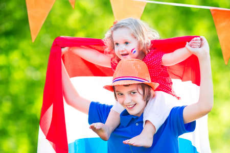Two Dutch children, teenager boy and funny little girl, celebrating national holiday of Netherlands playing in a garden decorated with Holland and Oranje flagsの写真素材
