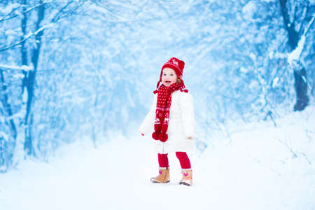 Happy laughing toddler girl wearing a white down jacket and red knitted hat and scarf playing and running in a beautiful snowy winter park on Christmas dayの写真素材