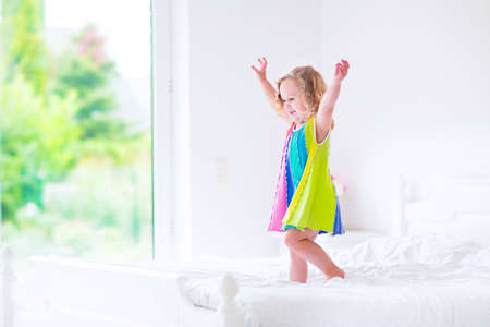 Cute little curly toddler girl in a colorful dress jumping on a big white bed laughing and having fun on a sunny weekend morning in a bedroomの写真素材
