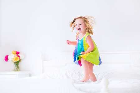 Cute little curly toddler girl in a colorful dress jumping on a big white bed laughing and having fun on a sunny weekend morning in a bedroomの写真素材