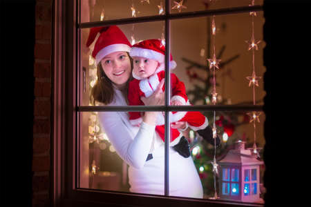 Young mother in red Christmas hat and her baby boy dressed in Santa costume standing next to a...