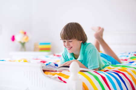 Happy smiling school boy reading a book relaxing on a white wooden bed with colorful blanket in a...