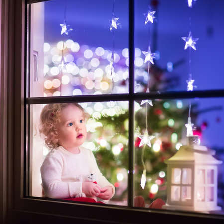 Cute curly toddler girl sitting with a toy bear at home during Xhristmas time, preparing to celebrate Xmas Eve, view through a window from outside into a decorated dining room with tree and lightsの写真素材