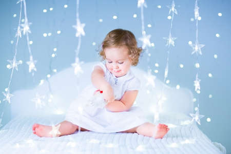 Adorable baby girl with curly hair playing with her bear between soft purple Christmas lightsの写真素材