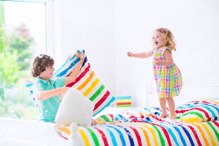 Two children, happy laughing boy and cute curly little girl having fun at pillow fight with feathers in the air jumping, laughing and giggling in a white bedroom with colorful bedding. Focus on jumping girl.の写真素材
