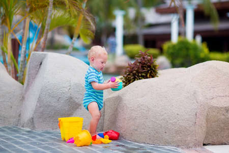 Cute little baby boy playing with plastic toy bucket and watering can at pool side in a tropical resort during summer vacationの写真素材