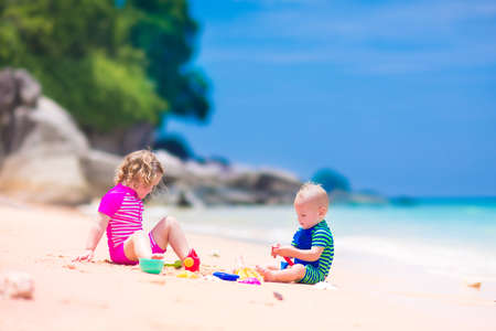 Happy baby boy and little curly toddler girl, brother and sister, playing with toy buckets and plastic shovel digging in sand on a beautiful exotic tropical beach with turquoise waterの写真素材