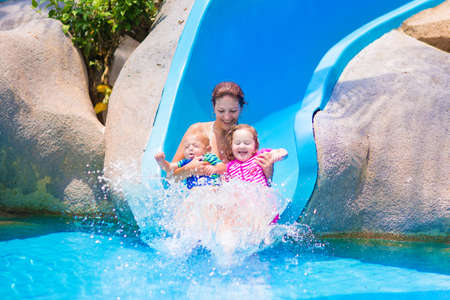 Young happy mother and two children, baby boy and toddler girl having fun at water sllide in a...