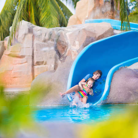 Happy boy and cute little girl, brother and sisterm having fun together on water slide in a swimming...