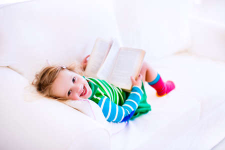 Happy laughing little girl reading a book relaxing on a white couch at homeの写真素材