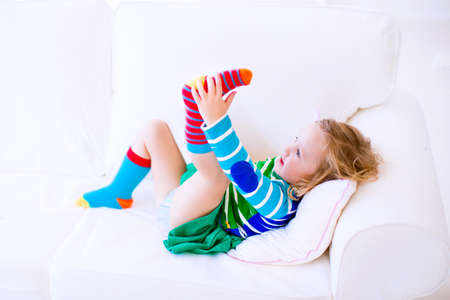 Funny little girl trying to put on warm socks on a white couch at homeの写真素材