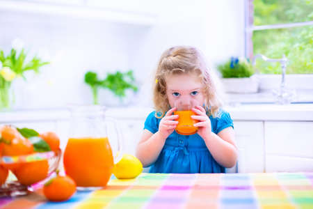 Cute funny little girl drinking freshly squeezed orange juice for healthy breakfast in a white kitchen with window on a sunny summer morningの写真素材