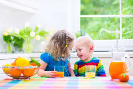 Cute funny little girl and adorable baby boy drinking freshly squeezed orange juice for healthy breakfast in a white kitchen with window on a sunny summer morningの写真素材