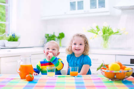 Cute funny little girl and adorable baby boy drinking freshly squeezed orange juice for healthy breakfast in a white kitchen with window on a sunny summer morningの写真素材