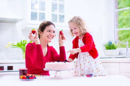 Happy family, young mother and her adorable little daughter, cute curly toddler girl in a red dress, baking fresh strawberry cream cake with fruit and berry in a white kitchen on a sunny summer dayの写真素材