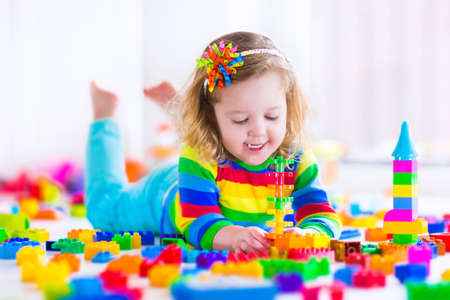 Cute funny preschooler little girl in a colorful shirt playing with construction toy blocks building a tower in a sunny kindergarten room Cute funny preschooler little girl in a colorful shirt playing with construction toy blocks building a tower in a sunny kindergarten room