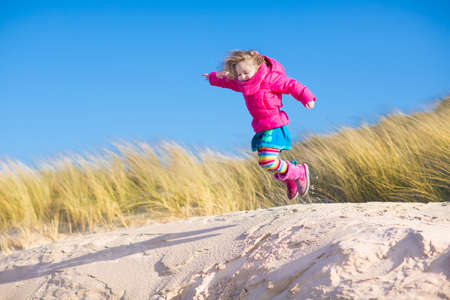 Happy funny little girl, adorable curly toddler, running and jumping in sand dunes enjoying family vacation at the North Sea, Holland, Netherlands on a sunny winter day at the beachの写真素材