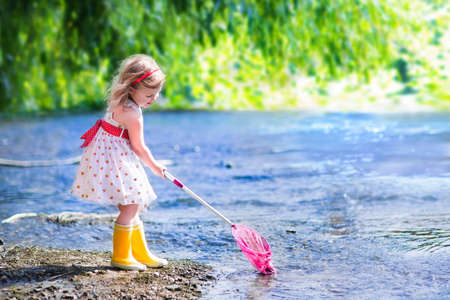 Child playing in a river. Cute little girl in a summer dress and rain boots catching fish and frog with a colorful net standing in water. Kids play outdoors. Young explorer and fisherman in wild nature.の写真素材