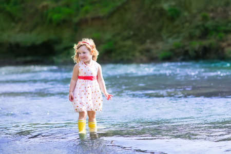 Adorable little curly toddler girl wearing a red and white dress playing at a river shore throwing...