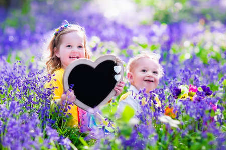 Children playing in bluebells forest. Little girl and baby boy holding a wooden heart shape chalk board standing in a park with beautiful spring bluebell flowers. Copy space for your text. Kids having fun outdoors in spring.の写真素材